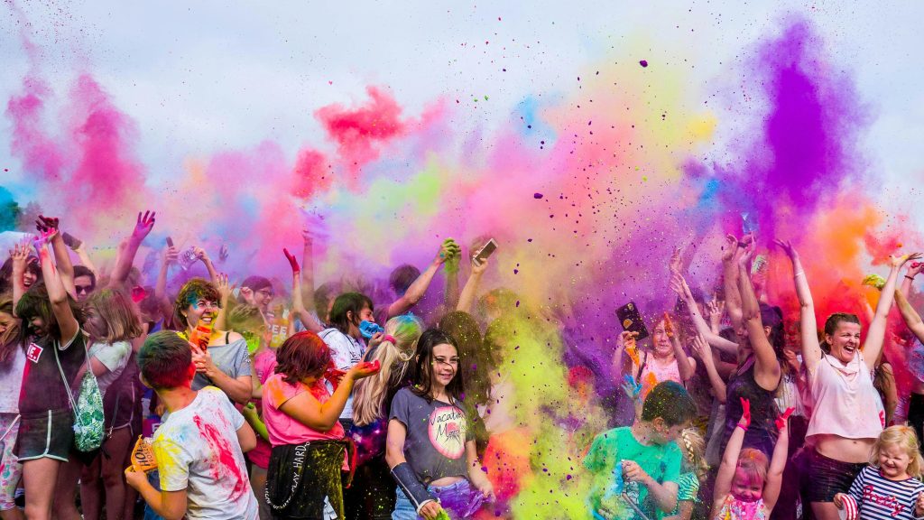 A joyful crowd covered in vivid colors celebrates a festival outdoors with flying powder.